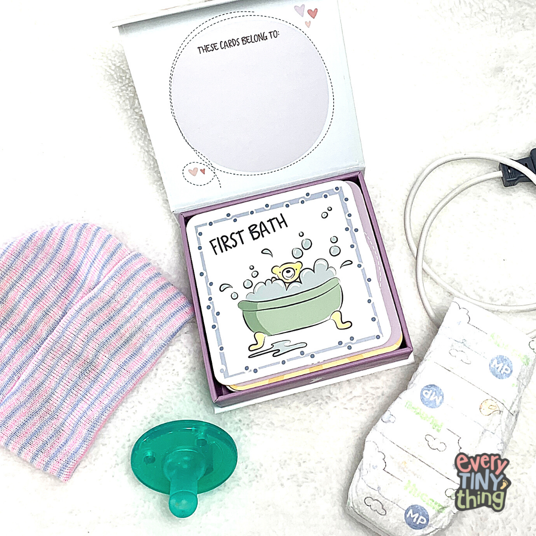 nicu milestone card box with "first bath" card at top of deck. Box lid says "these cards belong to" on the inside lid. Surrounded by newborn preemie baby hat, preemie pacifier, micropreemie diaper and NICU pulse-ox cord on white background with every tiny thing logo overlaid